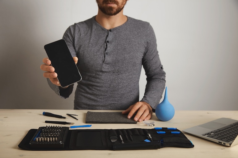 Bearded professional shows repaired fixed smartphone on camera after service replacement, above his specific tools in toolkit bag near laptop on wooden white table